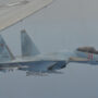 File Photo of Russian SU-35 fighter jet in flight, along with portion of wing of U.S. aircraft, adapted from image at defense.gov, with photo credit to U.S. Navy
