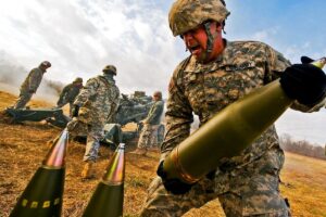 File Photo of Soldier Carrying 155mm Artillery Shell, with other Soldiers and Shells, and Artillery Piece, Nearby, adapted from army.mil image